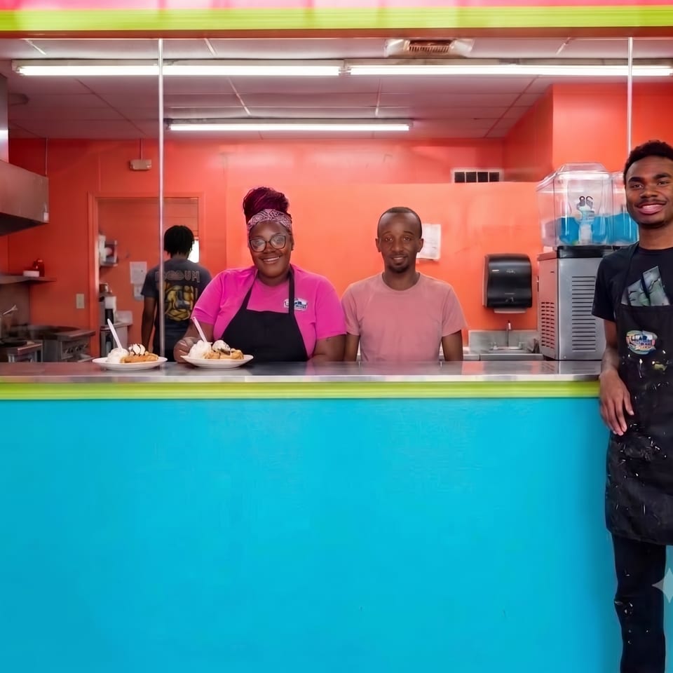 Three smiling employees stand behind a vibrant blue counter with two plates of dessert.