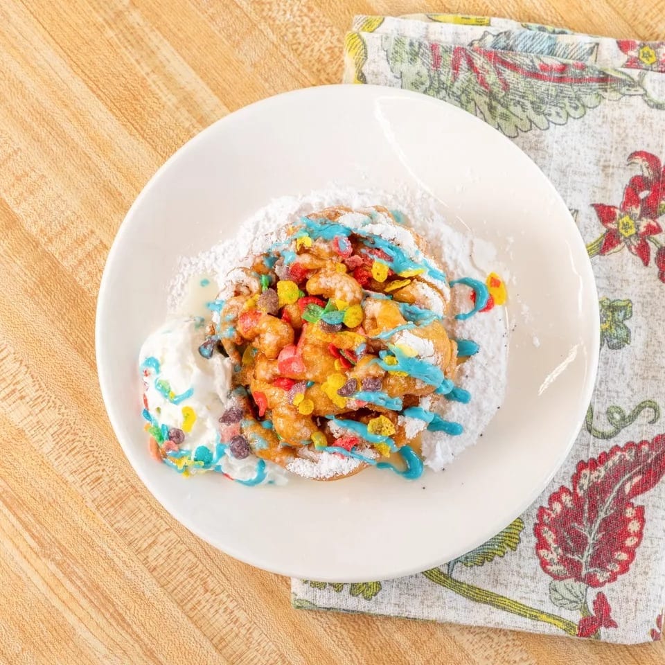 Funnel cake topped with colorful cereal, blue icing, powdered sugar, and whipped cream.