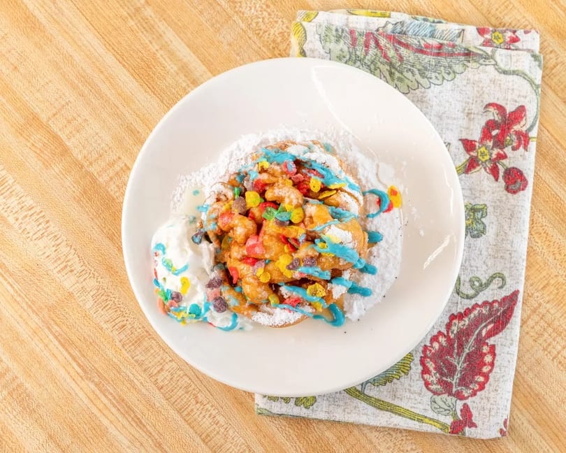 Funnel cake topped with colorful cereal, blue icing, powdered sugar, and whipped cream.