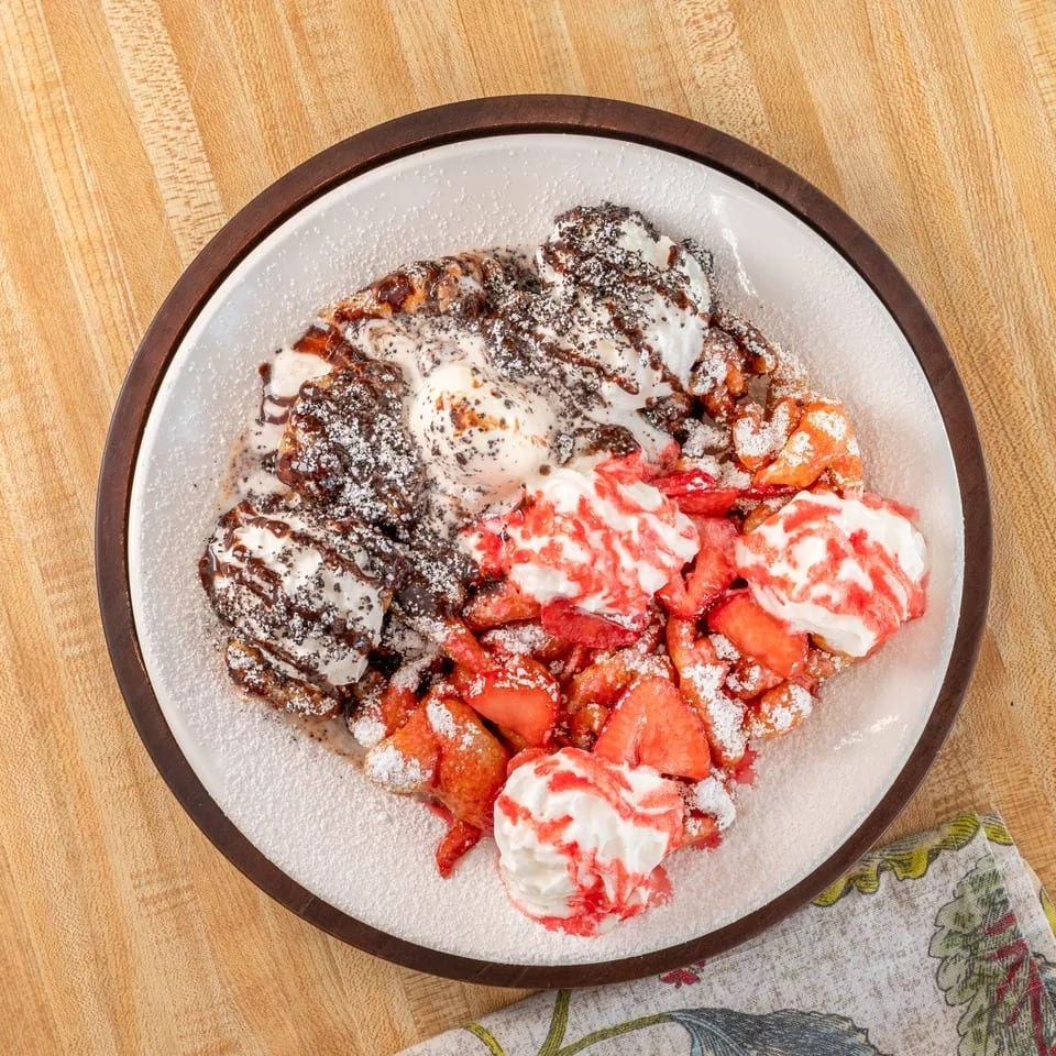 Funnel cake topped with strawberries, whipped cream, chocolate syrup, and powdered sugar.