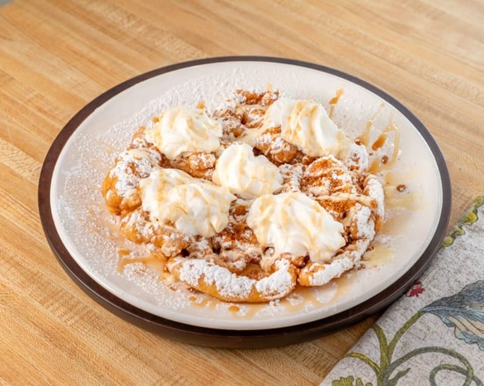 Funnel cake topped with whipped cream, powdered sugar, and a drizzle of syrup on a white plate.