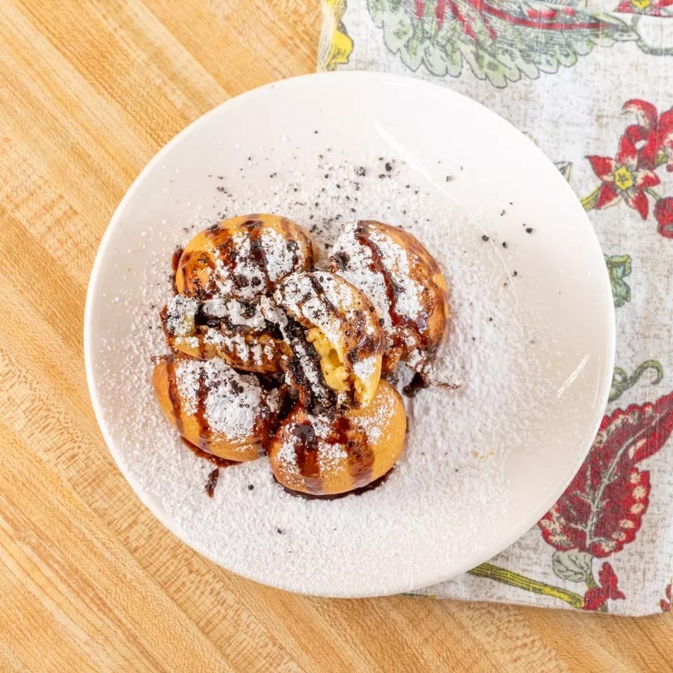 A white plate holds five golden-brown pastries drizzled with chocolate and powdered sugar.