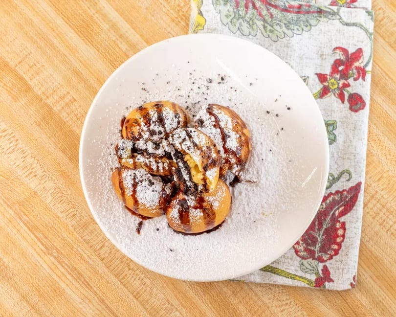 A white plate holds five golden-brown pastries drizzled with chocolate and powdered sugar.