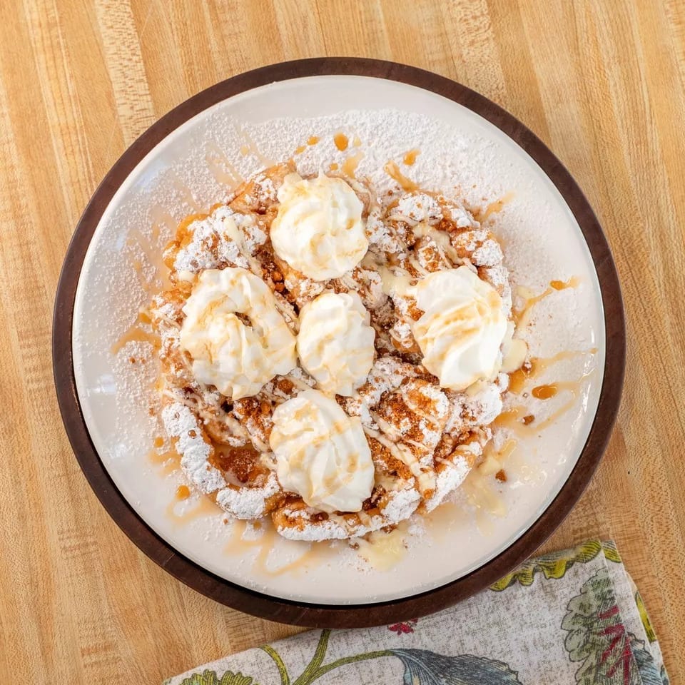 Funnel cake topped with whipped cream, powdered sugar, and caramel drizzle on a white plate.