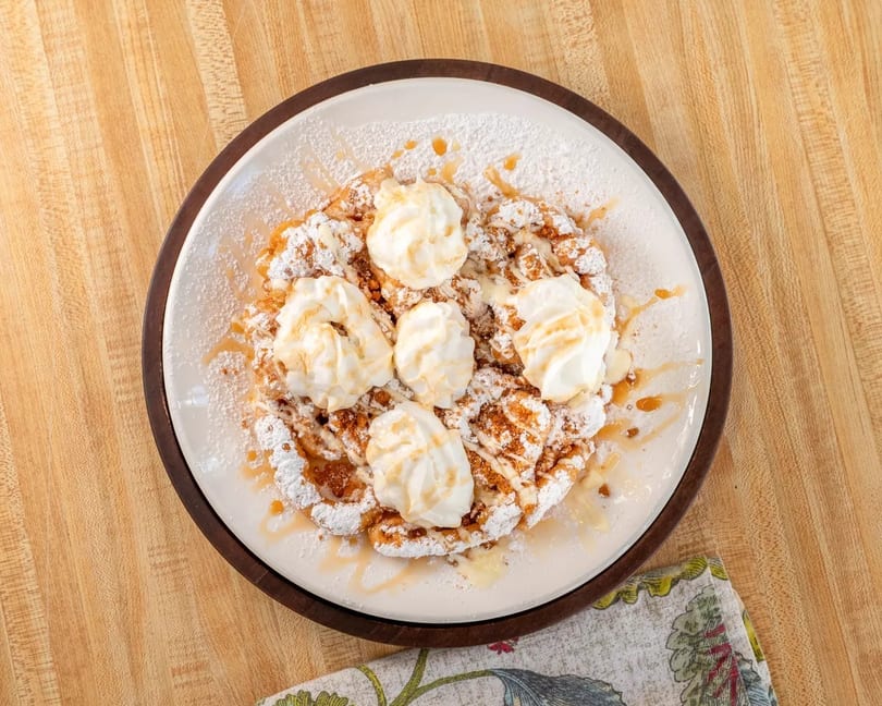 Funnel cake topped with whipped cream, powdered sugar, and caramel drizzle on a white plate.