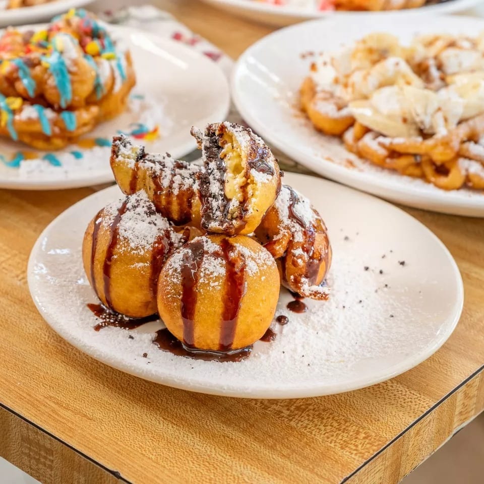 Plates of various fried desserts, including chocolate-filled dough balls and funnel cakes with toppings.