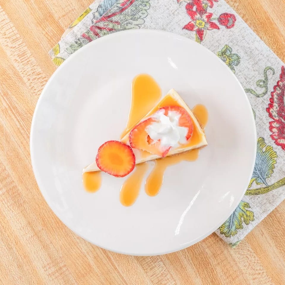 Cheesecake slice with caramel drizzle, whipped cream, and fresh strawberries on a white plate.