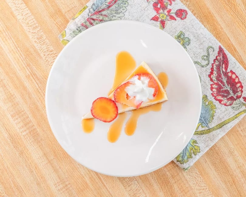 Cheesecake slice with caramel drizzle, whipped cream, and fresh strawberries on a white plate.